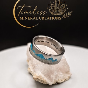 May include: A silver-toned metal ring featuring a mountain range inlay made of textured grey material and bright blue stone fragments. The interior of the band is engraved with the text "Gracie 2013-20". The ring rests on a rough, translucent white crystal.