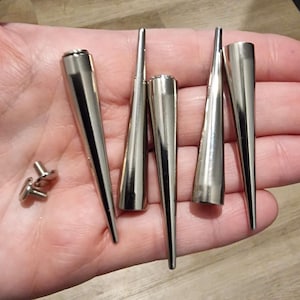 Op de afbeelding: Vijf zilveren metalen kegelvormige studs met een puntige punt. De studs zijn in een rij gerangschikt, met de punten naar rechts gericht. Twee kleinere zilveren metalen studs zijn ook zichtbaar in de afbeelding.