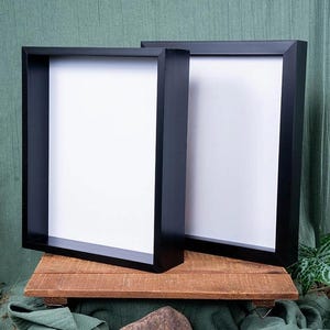 May include: Two black square picture frames with white mounts are displayed on a wooden surface. The frames are leaning against each other and are surrounded by green fabric and rocks.