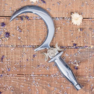 Może przedstawiać: Srebrny sierp księżycowy z symbolem potrójnego księżyca i wzorem pentagramu. Ostrze leży na drewnianej powierzchni z suszonymi kwiatami lawendy.