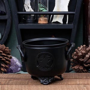 Op de afbeelding: Een zwarte ketel met een decoratief embleem, geplaatst op een houten oppervlak. De ketel heeft twee handvatten en staat op een decoratieve voet. Een zwarte plank met glazen potten en een bot is op de achtergrond. Het thema is gotisch of hekserig.