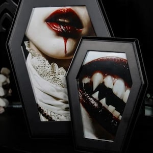 May include: Two black coffin-shaped picture frames with images of a vampire's mouth. The top frame shows a woman with red lipstick and blood dripping from her mouth. The bottom frame shows a close-up of a vampire's mouth with fangs.
