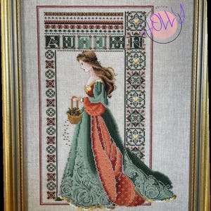 Può includere: Un ricamo a punto croce di una donna con un vestito verde e rosso che tiene un cesto di fiori. Il ricamo è incorniciato in una cornice dorata e la parola "Autunno" è ricamata sopra la donna.