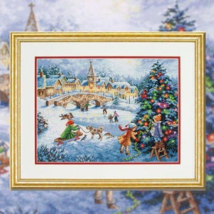 May include: A framed cross-stitch depicting a snowy winter scene. The artwork features a village with a bridge, people, and a decorated Christmas tree. The frame is gold-colored.