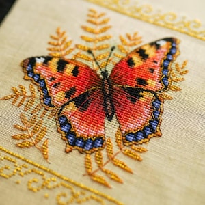 Puede incluir: Un detallado punto de cruz de una mariposa con alas rojas, naranjas, negras y azules, centrada en una tela beige. La mariposa está rodeada de hojas y bordes dorados bordados, mostrando el intrincado trabajo de aguja.