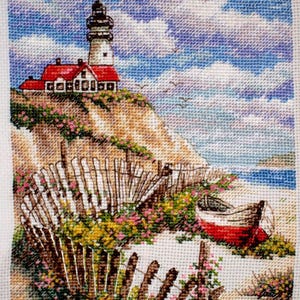May include: Cross-stitch of a coastal scene featuring a lighthouse with a red roof atop a cliff. A weathered fence and a small red and white boat are in the foreground. The sky is blue with fluffy white clouds, and the scene is filled with colorful flowers.
