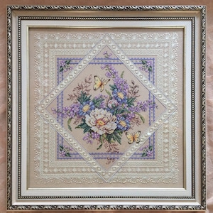 以下が含まれることがあります： 白いレースの縁取りで囲まれた、白い花と紫の花のブーケと2匹の黄色い蝶が描かれた、額装されたクロスステッチ刺繍です。