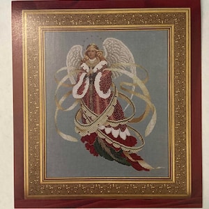 Op de afbeelding: Een ingelijst borduurwerk met kruissteek van een engel met een rode en witte jurk, omringd door een gouden lint. De engel heeft een halo en houdt een rol.