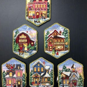 May include: Six hexagonal Christmas ornaments with a winter village theme. Each ornament features a different building, including a toy store, a bakery, a church, and a house. The ornaments are decorated with snow, trees, and lights.