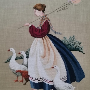 Puede incluir: Una mujer con un vestido rojo y azul con un delantal blanco camina con una bandada de gansos blancos. Lleva un palo en la mano. Los gansos la siguen en fila.