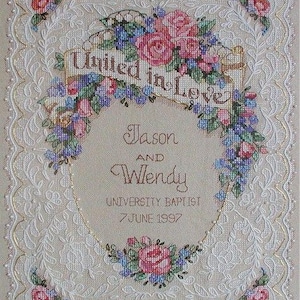 May include: A cross-stitch wedding sampler featuring the text "United in Love" above a floral design of pink and blue flowers. The names "Jason and Wendy" are embroidered below, with the date "7 June 1997". The sampler has a decorative lace border.