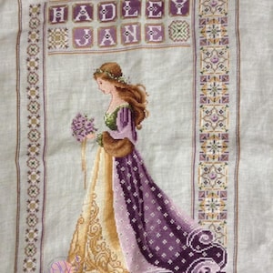 May include: Cross-stitch of a woman in a long, flowing gown, holding a bouquet. The dress is gold and purple, with intricate patterns. The name "HADLEY JANE" is stitched at the top. The background is a light beige.