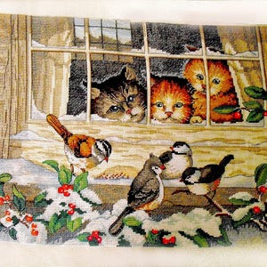 Puede incluir: Una escena de punto de aguja representa a tres gatos mirando por una ventana a pájaros en un paisaje nevado. Los gatos son atigrados marrones y naranjas. Los pájaros están posados en ramas cubiertas de nieve con bayas rojas. El marco de la ventana es de madera.