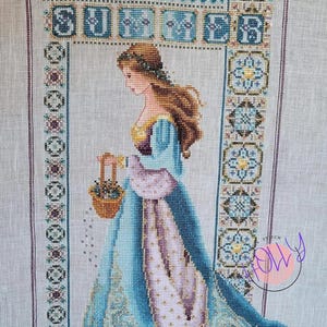 Può includere: Un ricamo a punto croce che rappresenta una donna con un vestito blu e viola che tiene un cesto di fiori. La donna è in piedi in una porta con un disegno floreale. Il ricamo è incorniciato da un bordo di fiori e motivi geometrici.