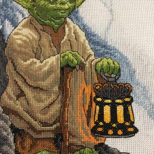 May include: A green and brown cross-stitch of Yoda from Star Wars holding a lantern and a walking stick. The background is a light blue and white.