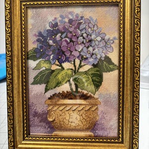 Può includere: Opera d'arte a punto croce incorniciata raffigurante una pianta di ortensia in un vaso decorativo. I fiori sono viola e blu, con foglie verdi. La cornice è dorata e ornata.