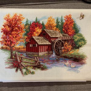 Può includere: Punto croce di una scena rurale autunnale con un mulino rosso e una ruota idraulica, circondato da alberi dai vivaci colori autunnali. Una staccionata in legno e due anatre sono in primo piano. Una spilla a forma di ape è attaccata in alto a destra.