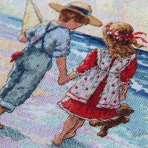 Peut inclure: Une broderie au point de croix représentant deux enfants, un garçon et une fille, se tenant la main en marchant sur une plage. Le garçon porte une chemise bleu et blanc à rayures, un jean bleu et un chapeau de paille. La fille porte une robe rouge et blanche à pois blancs. L'arrière-plan est un paysage marin bleu et blanc.
