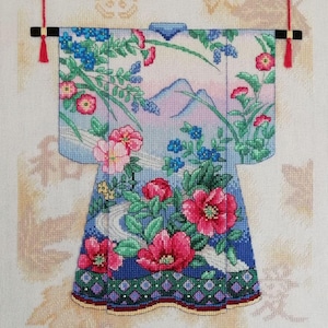 May include: A cross-stitch embroidery of a traditional Japanese kimono with a floral pattern and mountain landscape. The kimono is hanging on a wall with red tassels.