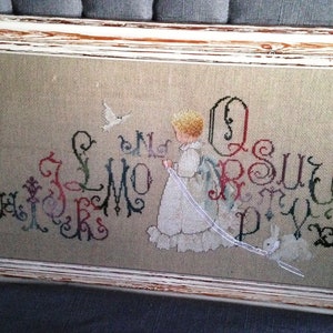 May include: A framed cross-stitch piece with a girl holding a string with a bird and two bunnies at the end. The cross-stitch features the alphabet in a decorative font with floral accents.