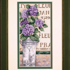 Puede incluir: Imagen de punto de cruz de hortensias moradas en un cubo galvanizado, enmarcada en verde. El fondo presenta texto como "FLEU" y "JAN 9729". El cubo tiene una corona y el número 3.