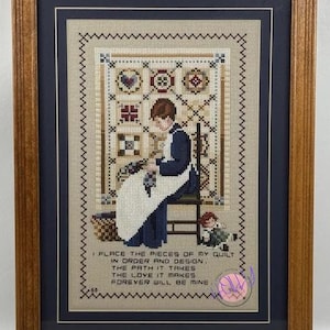 Può includere: Opera d'arte a punto croce incorniciata raffigurante una persona che cuce una trapunta, con una citazione sul quilting. Il pezzo presenta un design dettagliato con vari motivi e colori, incorniciato in legno.