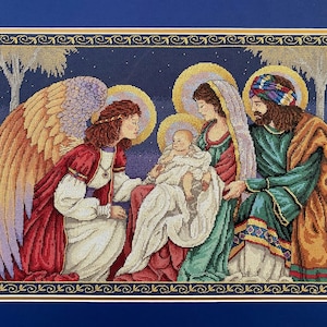 May include: A framed cross-stitch depicting a religious scene. An angel with large wings kneels before a baby held by a woman, with a man standing beside them. The figures are surrounded by halos, with a starry night background.