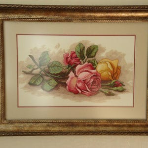 Puede incluir: Obra de arte en punto de cruz enmarcada con un ramo de rosas en tonos rosa y amarillo, con hojas verdes. La obra de arte está sobre un fondo neutro y enmarcada en un marco dorado y ornamentado.