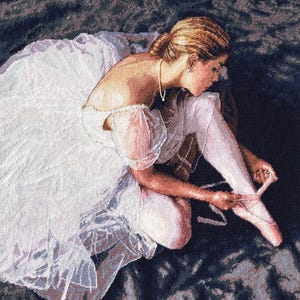 May include: A ballerina wearing a white tutu sits on a dark background, tying the ribbon on her pointe shoe.