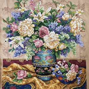 May include: A cross-stitch of a floral still life. A large vase is filled with lilies, peonies, and roses in pink, yellow, and purple. A smaller bowl of flowers sits beside the vase, on a gold fabric.