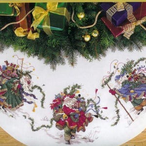 Peut inclure: Une jupe de sapin de Noël blanche avec un motif brodé vert, rouge et bleu de trois personnages portant des cadeaux et de la verdure. Les personnages sont entourés de vignes vertes et la jupe a un bord festonné.