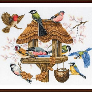 Op de afbeelding: Een vogelvoederhuisje met een rieten dak, omringd door verschillende kleurrijke vogels, waaronder blauwe mezen, een putter en een kardinaal. De voederbak is gevuld met zaden en noten.