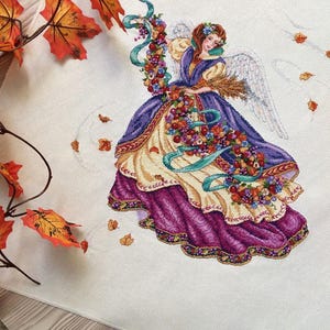 Pode incluir: Um bordado de ponto cruz que representa um anjo com um vestido roxo e dourado, segurando um buquê de folhas de outono e flores. O anjo tem uma auréola e asas brancas. O fundo é branco com folhas espalhadas.