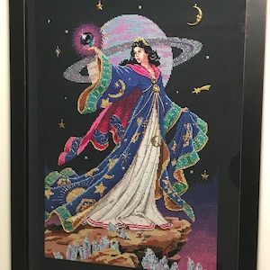 Könnte beinhalten: Eine gerahmte Kreuzstich-Stickerei, die eine Frau in einem blauen und goldenen Kleid zeigt, die eine leuchtende Kugel hält. Der Hintergrund ist dunkelblau mit Sternen, einer Mondsichel und einem Planeten mit Ringen.