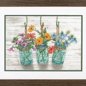 May include: Three glass jars with colorful flowers hanging from a white wooden wall. The jars are filled with pink, orange, and blue flowers.
