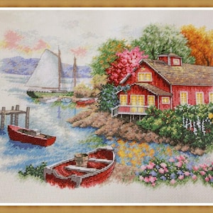 Può includere: Un paesaggio a punto croce raffigura una casa rossa vicino a un lago con una barca a vela e due barche a remi rosse. La scena comprende un molo, alberi colorati e fiori, il tutto incorniciato in un bordo di legno. L'opera è dettagliata e vibrante.