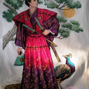 Puede incluir: Un bordado de punto de cruz que representa a un guerrero samurái con una túnica roja y morada, sosteniendo dos espadas y un abanico. Un pavo real se encuentra a la derecha del guerrero, con un plumaje verde y azul. El fondo es una tela blanca con un pino verde y un sol amarillo.