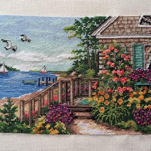 May include: A cross-stitch landscape depicts a coastal scene with a house, flowers, and water. The house has a brown roof and green shutters. Colorful flowers bloom around the house and along a wooden railing. Sailboats and seagulls are visible in the blue water.