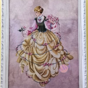 Può includere: Un ricamo a punto croce di una donna con un vestito giallo decorato con rose e viti rosa. Tiene un mazzo di rose rosa ed è circondata da una ghirlanda di rose.