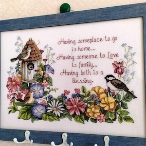 May include: A framed cross-stitch artwork with a blue wooden frame. The artwork features a birdhouse, birds, and colorful flowers. Text reads: "Having someplace to go is home... Having someone to Love is family... Having both is a blessing."
