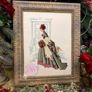 Op de afbeelding: Een ingelijst kruissteekpatroon van een vrouw in een groene jurk en een witte sjaal die een mand met hulst vasthoudt. De lijst is versierd en goudkleurig met een kruissteekontwerp van het woord "Holly" in de rechterbenedenhoek.