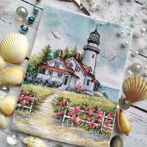 May include: A cross-stitch of a coastal scene featuring a white lighthouse with a black top, a red-roofed house, and a path lined with pink flowers. The artwork is surrounded by seashells, clear glass beads, and gold scissors.