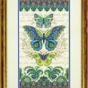 Op de afbeelding: Ingelijste kruissteekkunst met drie kleurrijke vlinders in blauw, groen en geel. De vlinders staan tegen een crèmekleurige achtergrond met tekst in scriptstijl en decoratieve randen. De lijst is goudkleurig.
