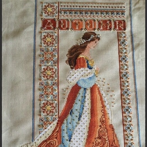 Può includere: Ricamo a punto croce di una donna con un vestito blu e arancione con la parola "AUTUMN" sopra di lei. Il ricamo è incorniciato da un bordo di perline.