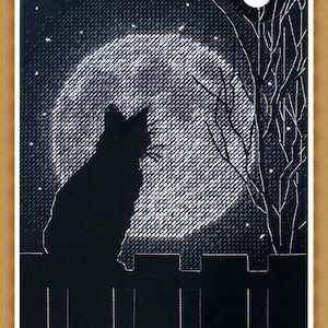 May include: A black cat silhouette sits on a fence, with a full moon and bare tree branches in the background. The artwork is framed in a light brown wooden frame. The night sky is filled with stars.