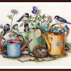 May include: Cross-stitch artwork featuring three watering cans in blue, green, and yellow, adorned with butterflies, birds, and daisies. A small bird's nest with blue eggs is also present. The artwork is framed in a brown wooden frame.