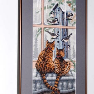 Può includere: Opera d'arte a punto croce incorniciata raffigurante due gatti con pelliccia arancione e marrone, che guardano fuori da una finestra uccelli e una casetta per uccelli. La scena è ambientata in inverno, con neve e ghiaccioli visibili.