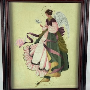 Può includere: Un ricamo a punto croce incorniciato che raffigura un angelo con una colomba bianca in mano. L'angelo indossa un vestito rosa e verde con una fascia viola e un'aureola bianca. Lo sfondo è giallo chiaro.