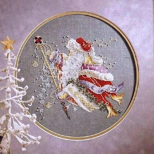 May include: A circular cross-stitch embroidery featuring a stylised figure in a red and white robe holding a tall staff with a square topper, accented by scattered beads on a grey fabric background, framed in a simple round wooden hoop.
