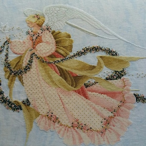 Puede incluir: Una bordado de punto de cruz que representa a un ángel con un vestido rosa y dorado, alas blancas y una guirnalda floral. El ángel sostiene un pequeño pájaro en su mano.